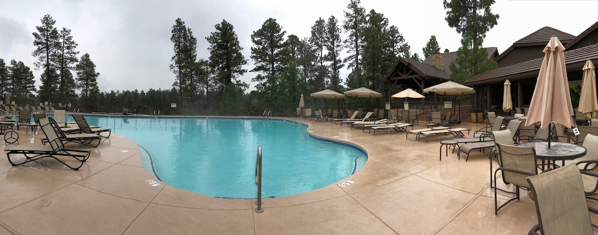 A spacious outdoor pool is surrounded by a large deck, featuring several sun loungers and umbrellas. Towering trees provide a natural backdrop, while the pool's clear water reflects the overcast sky.