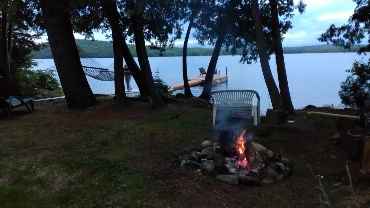 A fire pit surrounded by stones is lit in a lakeside setting, with a white chair positioned nearby. In the background, a wooden dock extends into the water, and a hammock is visible strung between trees. Scenic views of the lake and hills are present.