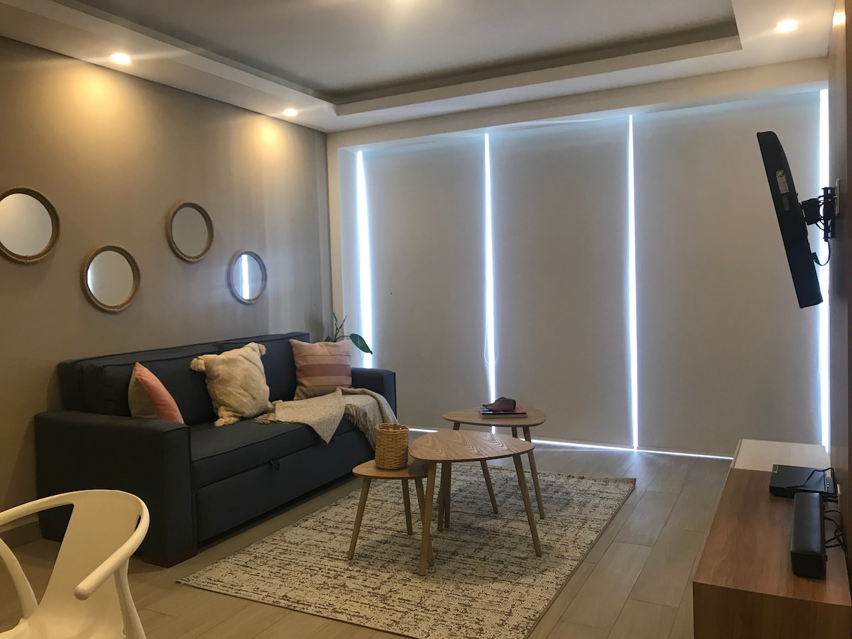 The living area is designed with a comfortable dark sofa accompanied by decorative pillows. Two wooden tables are positioned on a light-colored rug. Natural light filters in through sheer window coverings, creating a calm and welcoming space. Wall mirrors add visual interest.