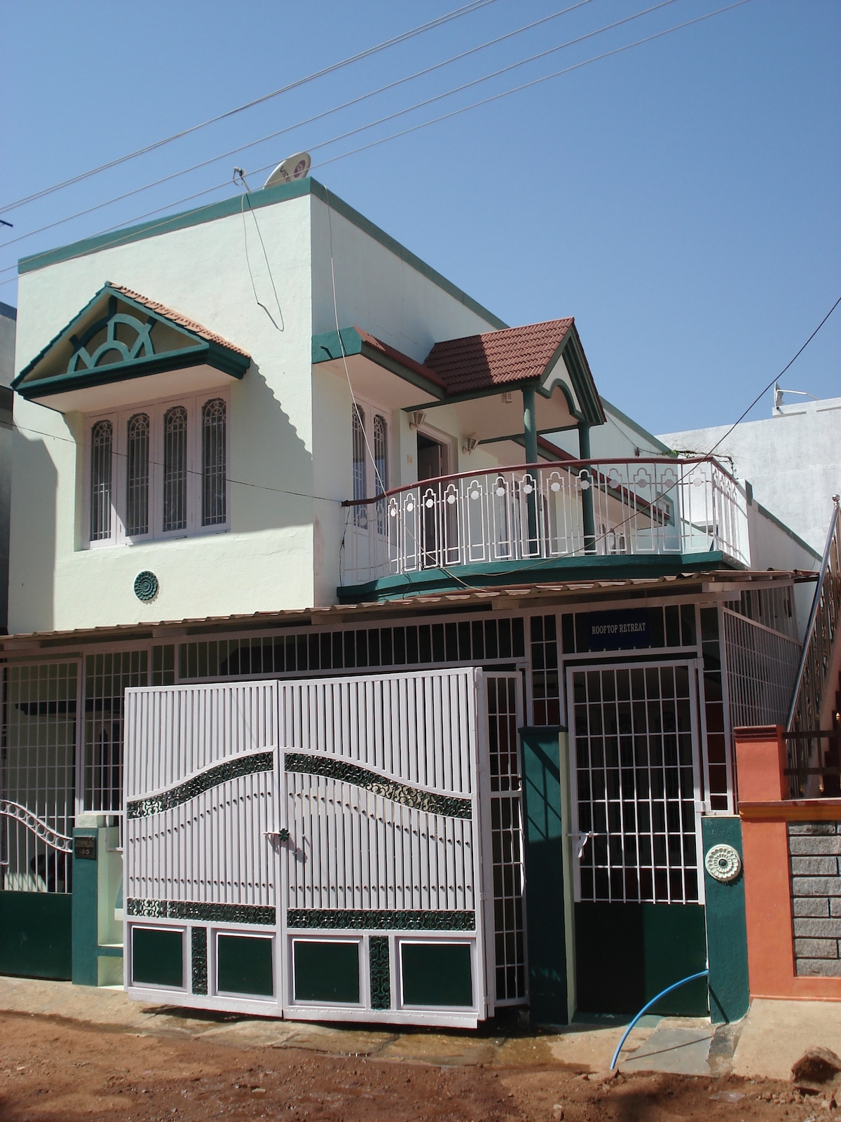The exterior of the house showcases a light green façade with decorative details. A white gate opens to a private entrance. A balcony with railings is visible on the first floor. The structure is set against a clear blue sky, emphasizing its inviting appearance.