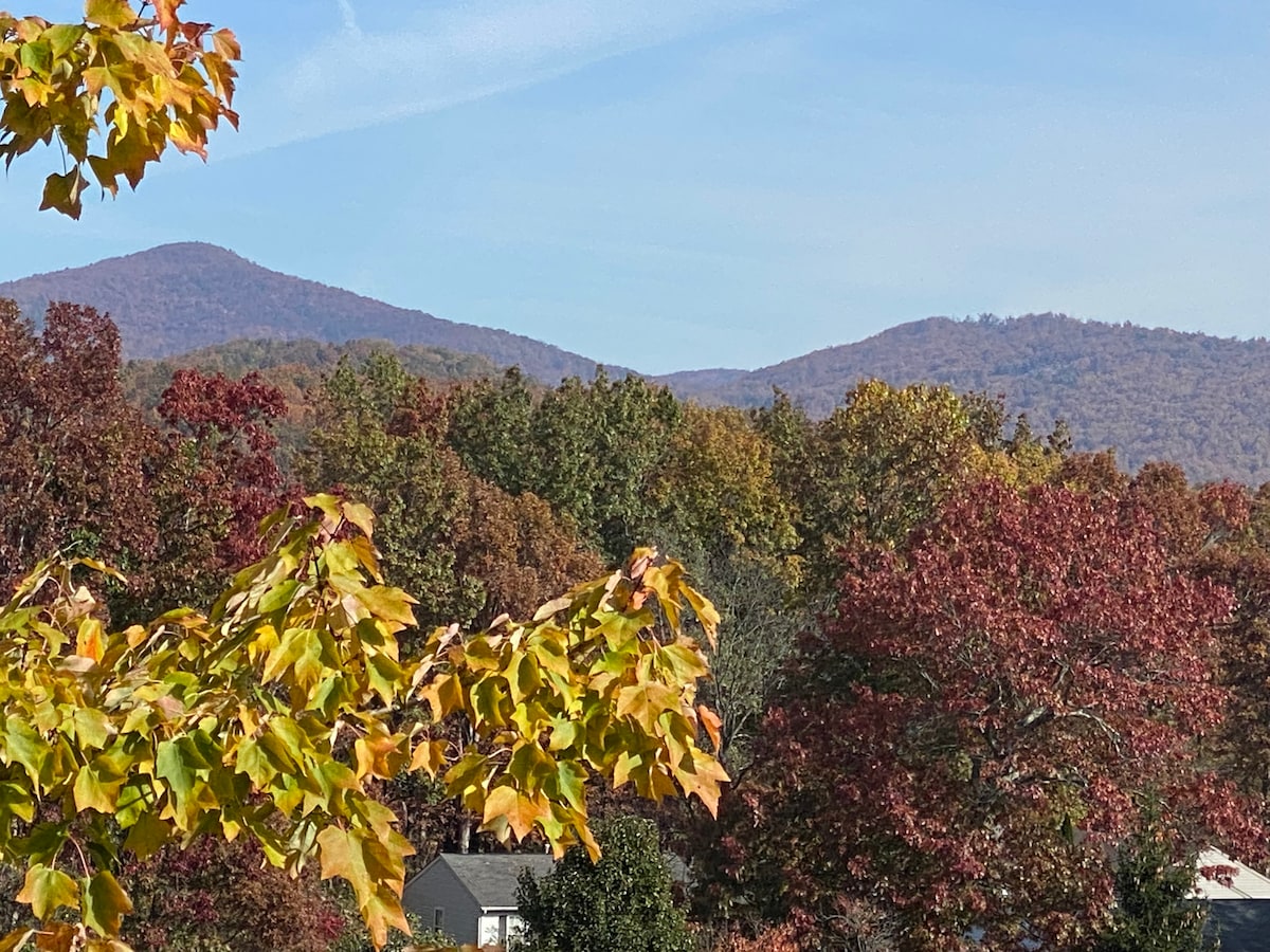 Early autumn foliage displays a rich tapestry of reds and greens, with distant mountains serving as a backdrop under a clear blue sky. The vibrant leaves gently frame the scene, emphasizing the natural beauty of the area.