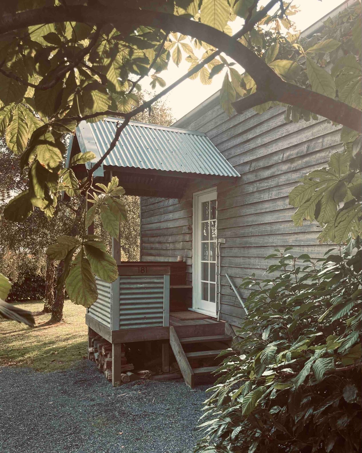 A wooden exterior is framed by lush greenery, with a covered porch leading to a white door. Stairs ascend from the gravel path to the entrance, and the corrugated metal roof provides shelter, blending harmoniously with the rustic barn structure.
