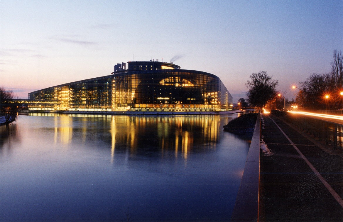 A modern building is reflected in the calm waters of the river, illuminated by soft lights as dusk falls. Smooth pathways alongside the water are visible, along with faint outlines of trees and street lamps providing gentle ambiance.
