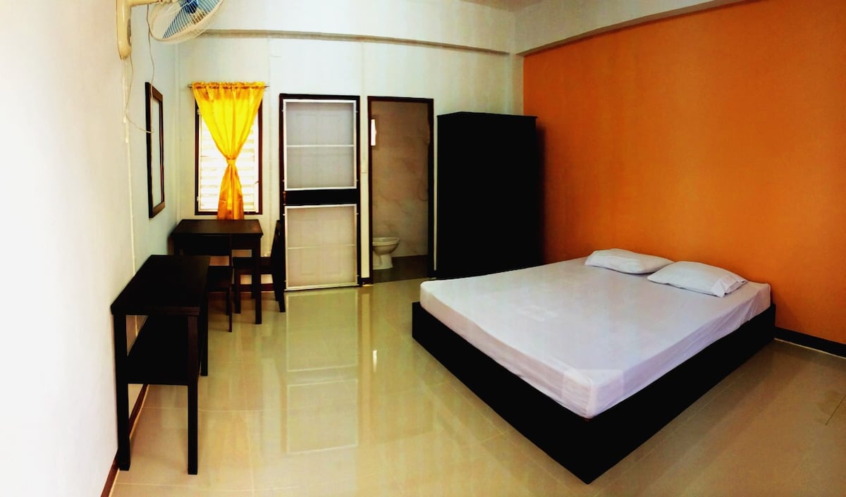 A spacious room is furnished with a king-sized bed and equipped with two black tables. Natural light enters through a window adorned with bright yellow curtains. A bathroom is visible in the background, complemented by an orange accent wall.