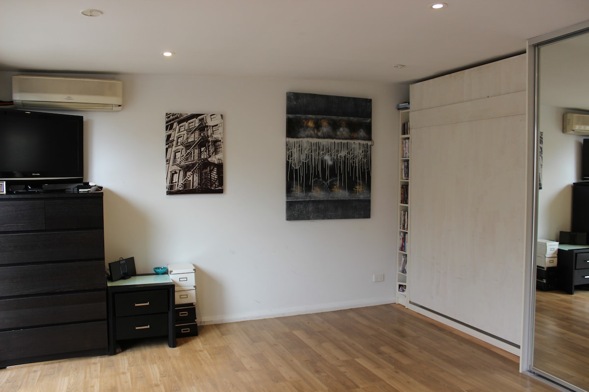 A studio space features a large mirrored wardrobe reflecting the light-colored walls. Stylish artwork adorns the walls, and a flat-screen television is mounted above a dark chest of drawers. Natural light fills the area, creating an open and airy environment.