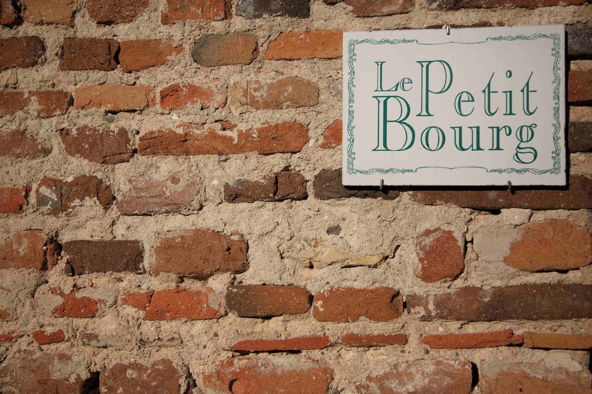 A rustic brick wall showcases a weathered sign reading 'Le Petit Bourg.' The texture of the bricks is prominently displayed, highlighting the earthy tones and aged appearance of the wall.