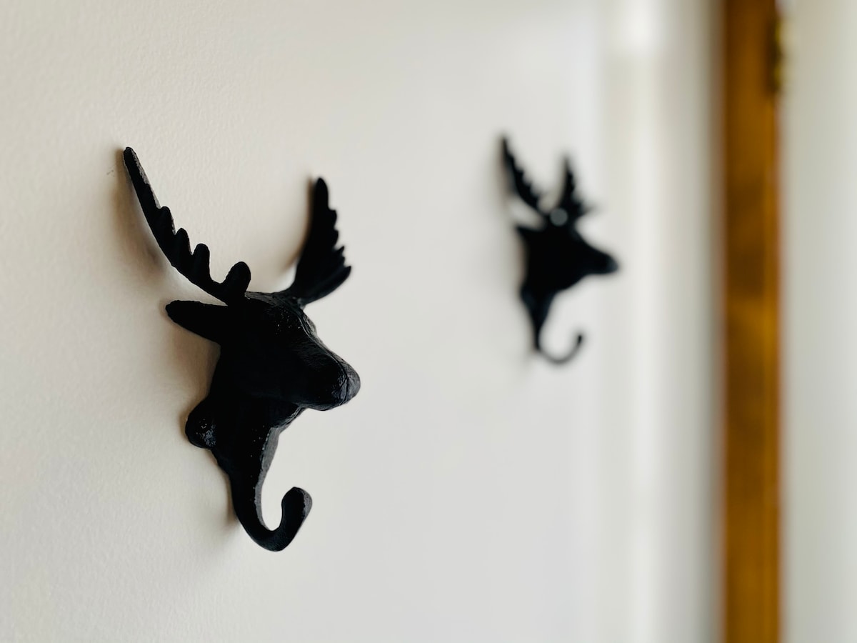 Two decorative black hooks shaped like moose heads are mounted on a white wall. Each hook features prominent antlers and a curved accent at the base, providing functional charm to the space.