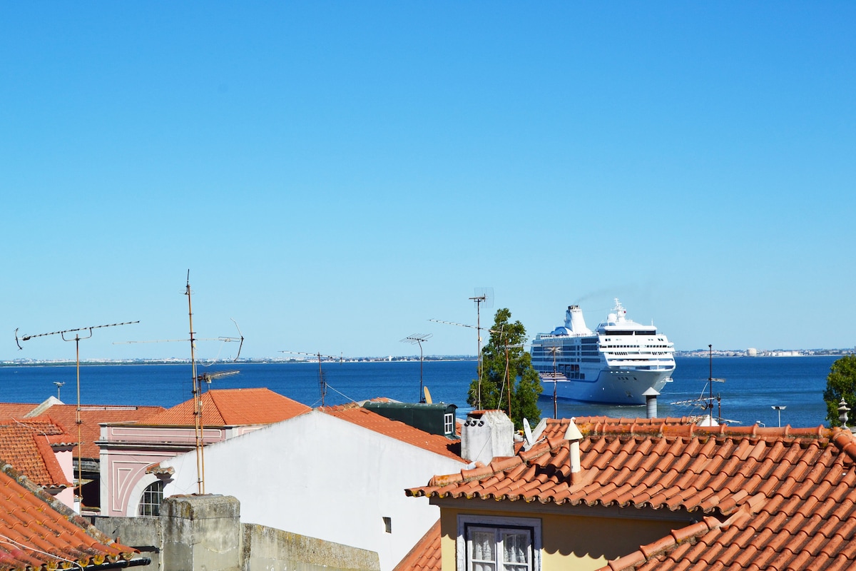 The image showcases a bright blue sky above the Tejo River, framed by traditional Portuguese rooftops with terracotta tiles. A large cruise ship is seen docked in the water, with distant land visible along the horizon, inviting a sense of calm and openness.
