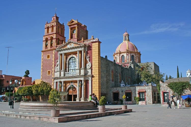 A vibrant town square features an ornate stone fountain surrounded by trees and walking paths. Historic buildings, showcasing a mix of colorful facades and architectural styles, dominate the scene, with towers and a dome rising against a clear blue sky.