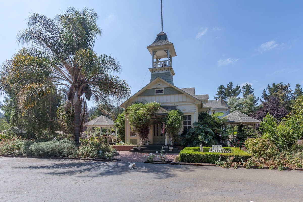 A charming exterior of the building is showcased, featuring a distinctive tower and lush landscaping. Tropical palms and various green plants enhance the inviting entrance, while a well-manicured pathway leads to the front porch. The setting reflects a tranquil country atmosphere.