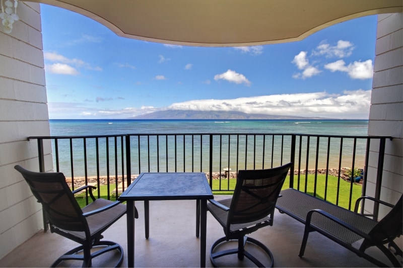 A private balcony overlooks the expansive ocean, with views of the island of Molokai in the distance. Two modern chairs are positioned around a small table, complementing the open and airy atmosphere created by large sliding doors.
