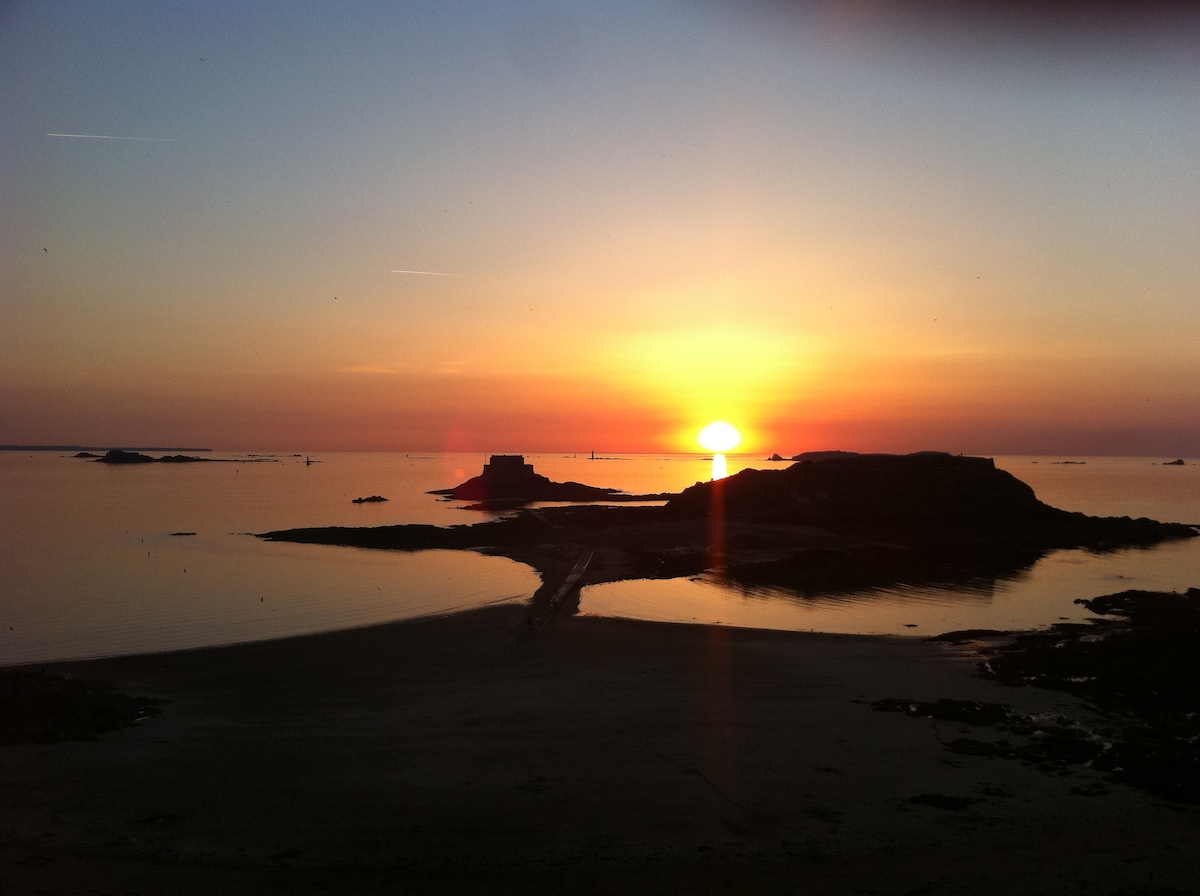 A serene coastal view captures a sunset over calm waters, with silhouettes of distant rocks reflecting the warm orange and yellow hues. The shoreline is visible, providing a tranquil setting as day transitions to night.