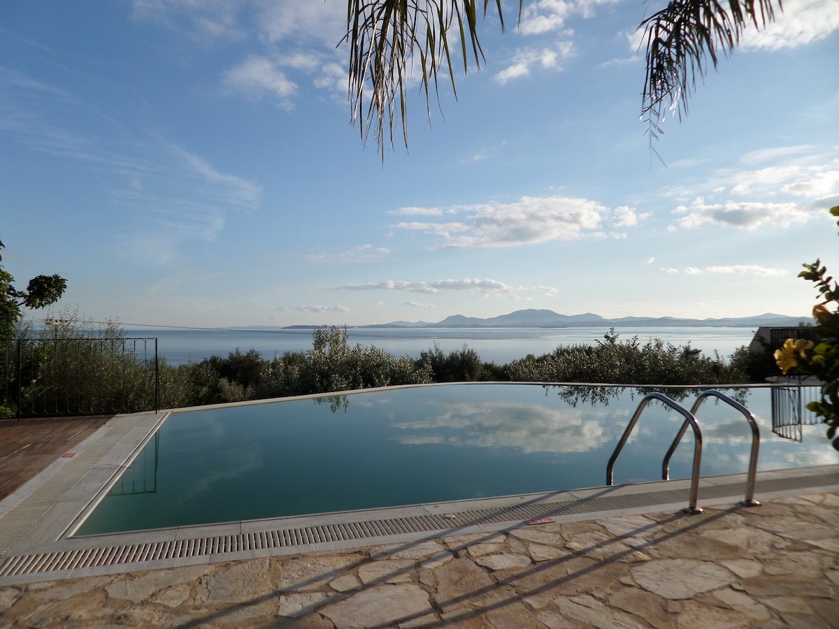 An extra long pool is visible, reflecting the clear blue sky and surrounding olive grove. The pool area is framed by lush greenery, with gentle waves of the sea in the background. This tranquil outdoor space offers stunning views of the coastline.