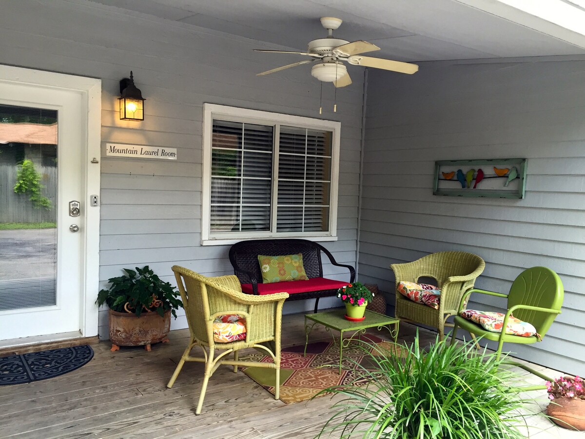 The covered wrap-around porch features a seating area with a black sofa and colorful chairs. A small table sits in the center, adorned with a potted flower. The space is complemented by a ceiling fan and a wall-mounted artwork depicting birds.