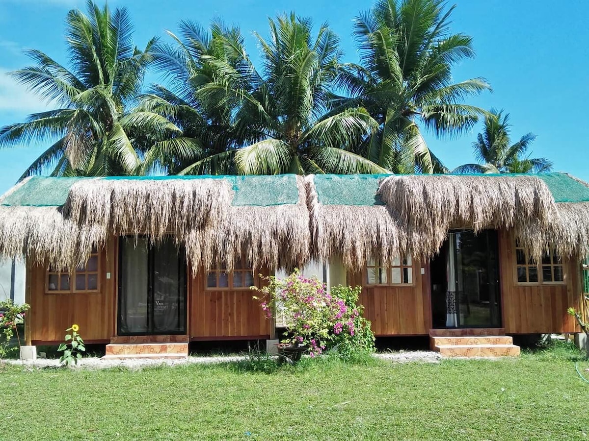 A cluster of wooden cabins is presented, each featuring thatched roofs. Lush green grass surrounds the cabins, and vibrant flowers bloom along the walkway. Towering palm trees provide shade, while a clear blue sky is visible above.