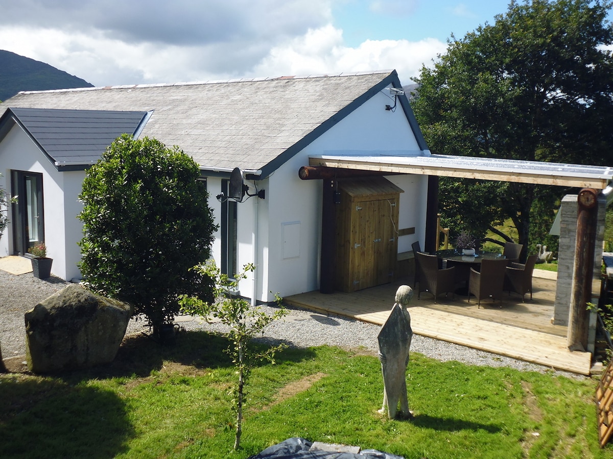 The exterior of the property features a modern, single-story structure with a mix of white walls and a gray roof. A covered wooden patio extends from the building, furnished with a dining table and chairs. Lush greenery surrounds the entrance and patio area.