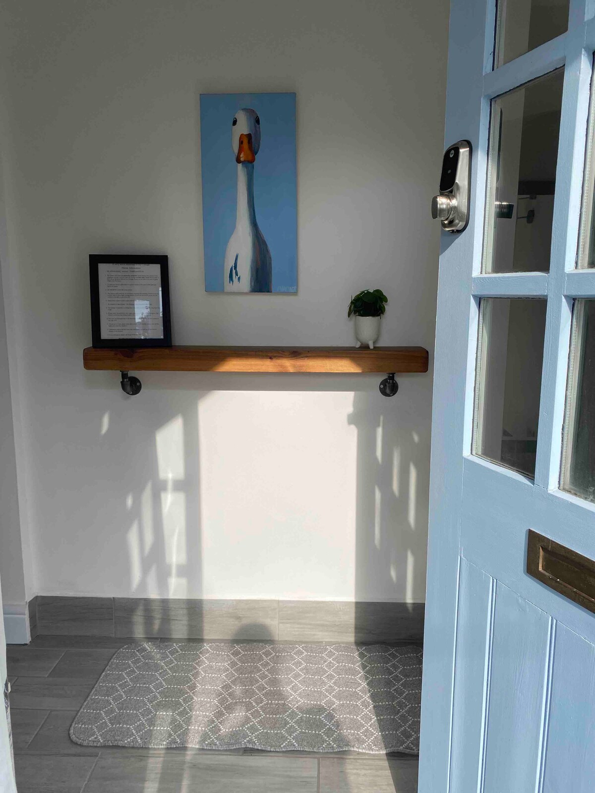 A bright entrance hall is illuminated by natural light, featuring a pale blue door. A wooden shelf is mounted on the wall above a small potted plant and a framed information sheet. A patterned mat is placed on the tiled floor.
