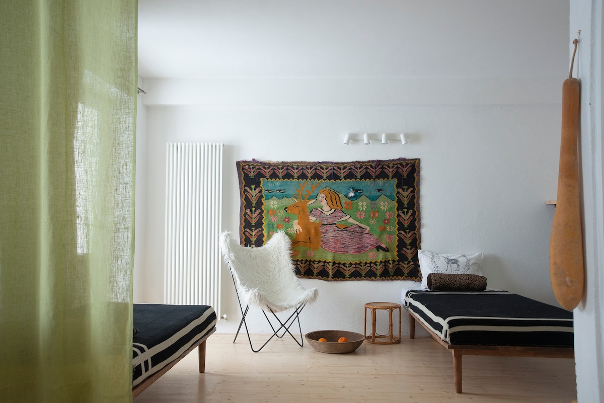 A cozy studio layout features two spacious beds with striped linens, positioned near a colorful wall tapestry. A white accent chair is situated next to a wooden stool, and a bowl of oranges adds a touch of warmth. Natural light fills the room through a large window.
