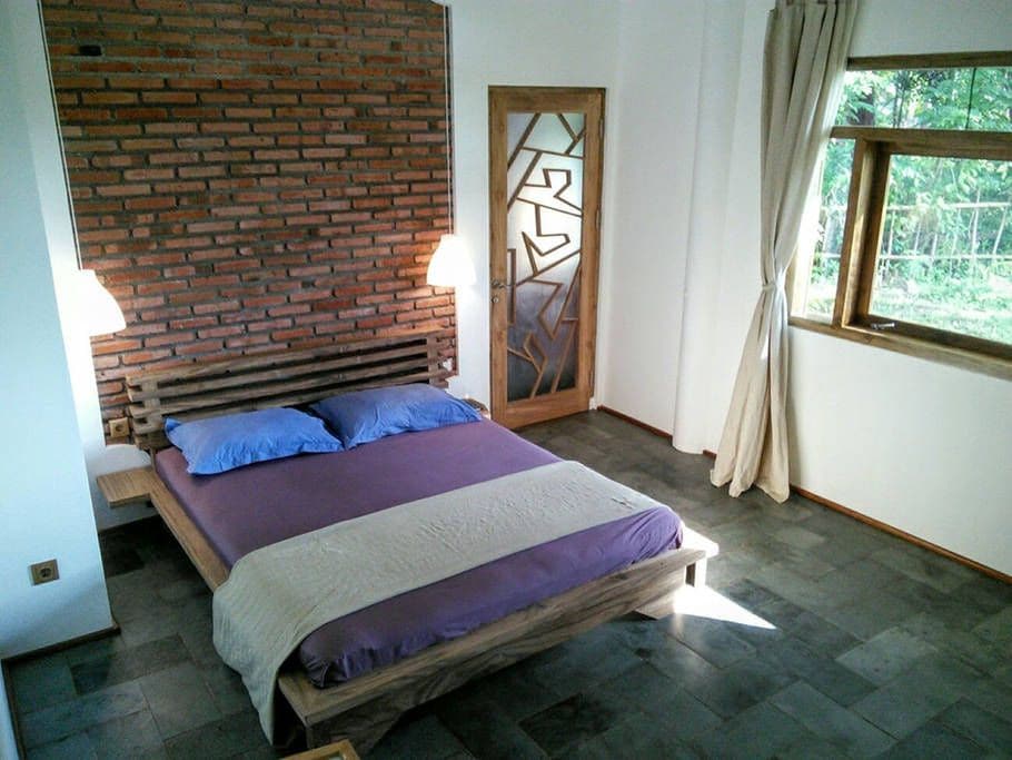 The room features a king-size bed with blue pillows and a soft beige blanket. A textured brick wall serves as a backdrop, while two bedside lamps provide soft lighting. Large windows invite natural light and offer views of the surrounding greenery.