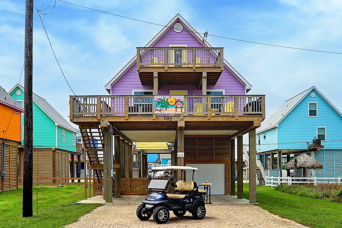 A colorful beach house stands elevated on stilts, featuring a spacious deck with multiple seating areas. A golf cart is parked nearby on the gravel, while vibrant hues of purple, blue, and orange define the neighboring buildings. Lush green grass surrounds the property.