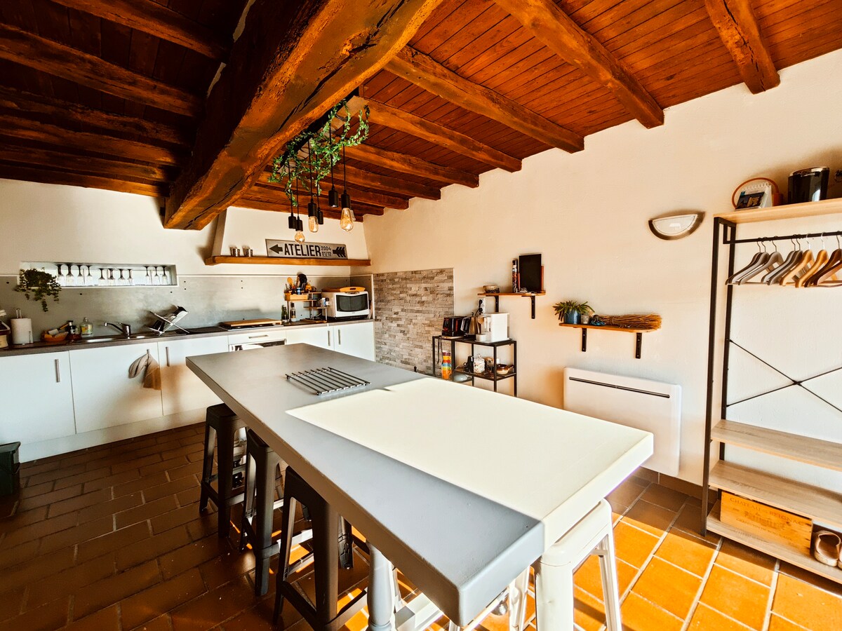 A spacious kitchen features a large central island with bar stools, surrounded by white cabinetry and rustic wooden beams overhead. Essential kitchen appliances are present, with a grey backsplash adding a modern touch. Natural light fills the room, highlighting the open layout.