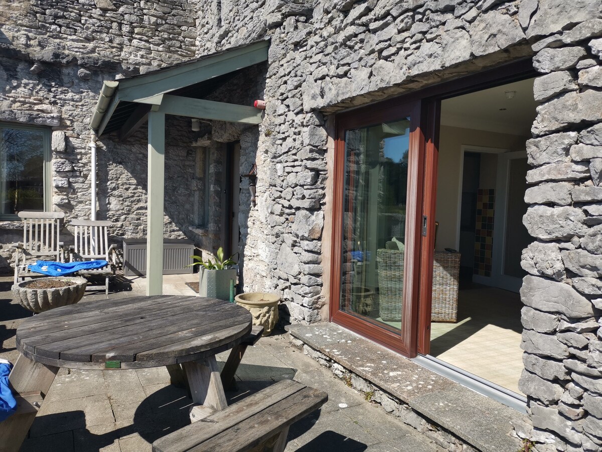 An outdoor patio area showcases a round wooden table with benches, surrounded by natural stone walls. Large glass doors open onto an interior space, allowing for seamless transition between indoor and outdoor areas. Sunlight highlights the textured stone exterior.