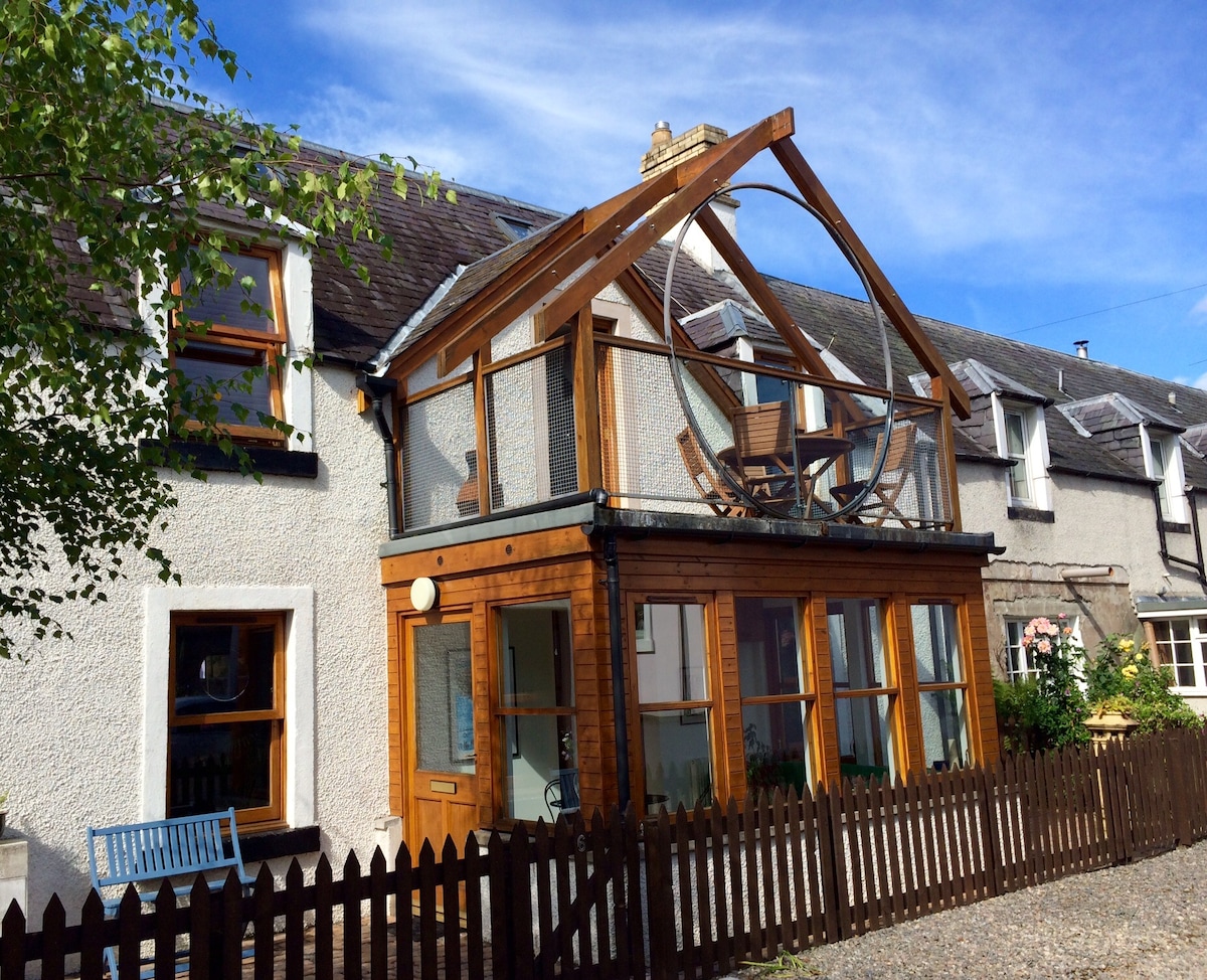 The cottage features a charming wooden garden room with large glass windows, allowing plenty of natural light. A circular seating arrangement is visible above. The exterior is complemented by a wooden fence and a blue bench, set against a clear blue sky.