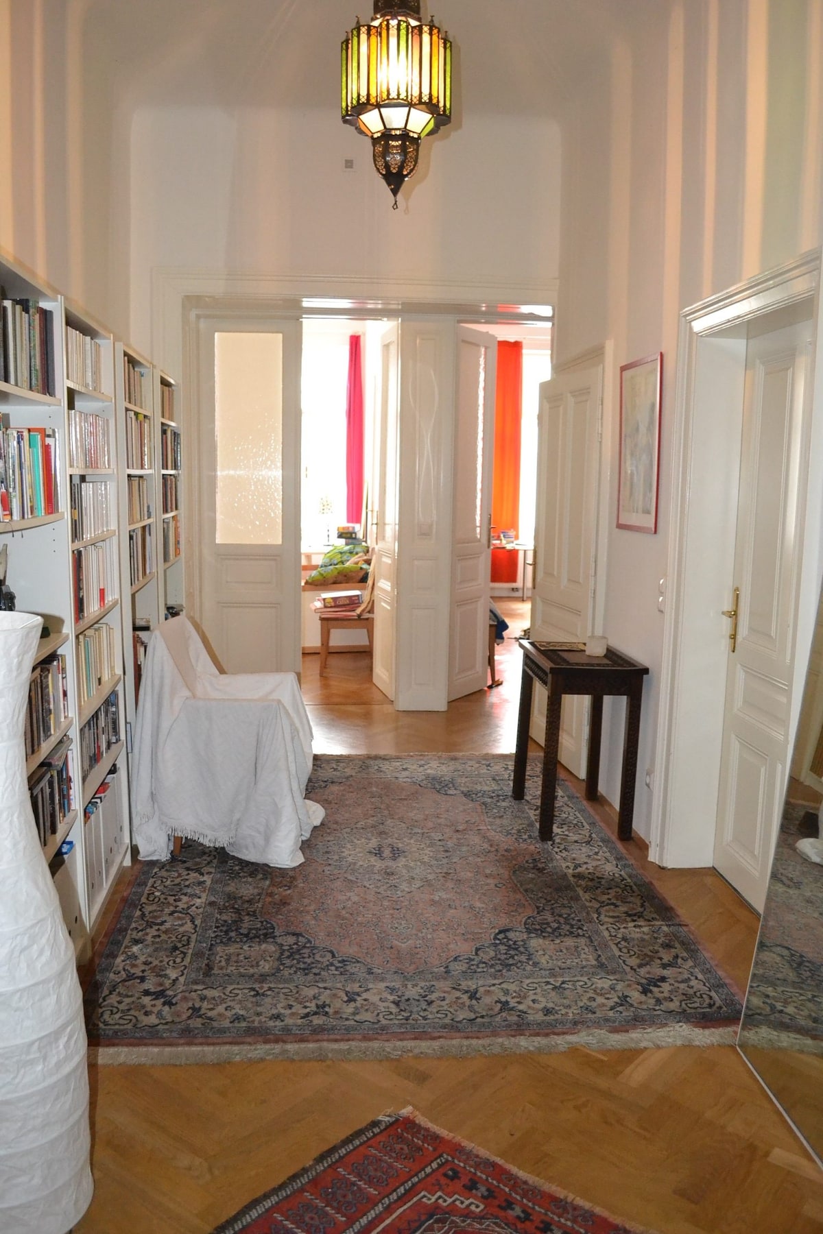 A hallway is presented with warm wooden flooring and a patterned area rug. Bookshelves line one side, filled with various books. Dual doors lead to a bright room beyond, where soft natural light filters through. A small table and a draped chair are also visible.