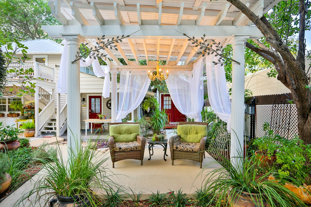 An outdoor seating area is featured under a white trellis adorned with sheer white curtains. Two comfortable chairs with green cushions are positioned beside a small glass table, surrounded by lush plants and a tranquil garden setting.