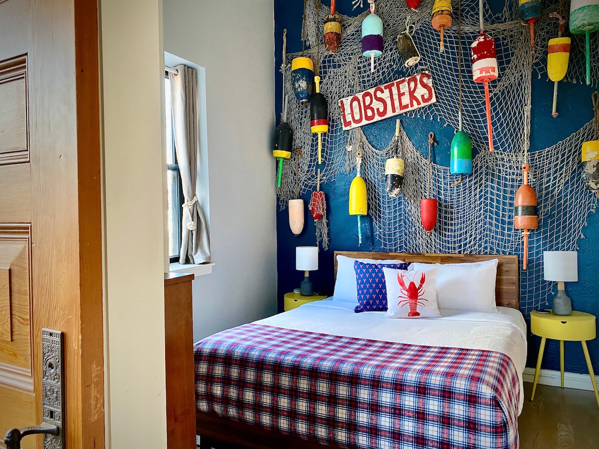 The Lobster Room features a spacious bed adorned with a patterned blanket. A vibrant display of colorful buoys is showcased against a deep blue wall, accompanied by fishing net decor. Light-colored nightstands and a unique lobster-themed pillow add character to the room.