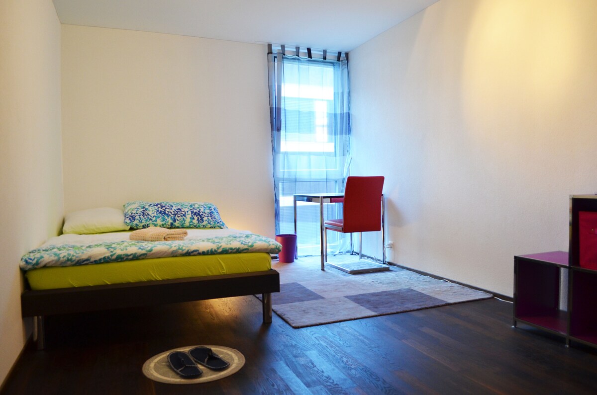 A private room features a spacious bed with colorful bedding and a design table positioned beside a window with sheer curtains. A light-colored area rug is placed on a dark wooden floor, creating a contrast in the inviting space.