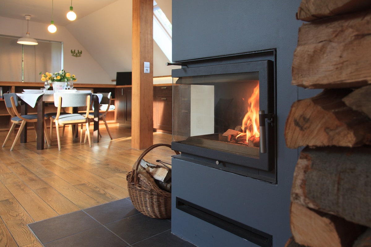 A modern fireplace with a glass front is prominently displayed, showcasing an inviting fire. A basket of firewood sits nearby. The background reveals a dining area with a table set for meals and wooden chairs, complemented by warm natural light streaming through windows.