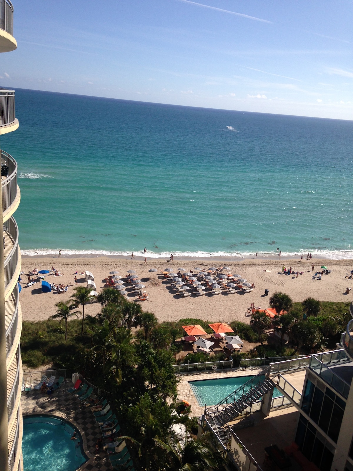An expansive beachfront view showcases a sandy shore lined with sun loungers and umbrellas. The distant horizon meets a shimmering turquoise sea under a clear blue sky. In the foreground, glimpses of a pool area surrounded by palm trees can be seen.