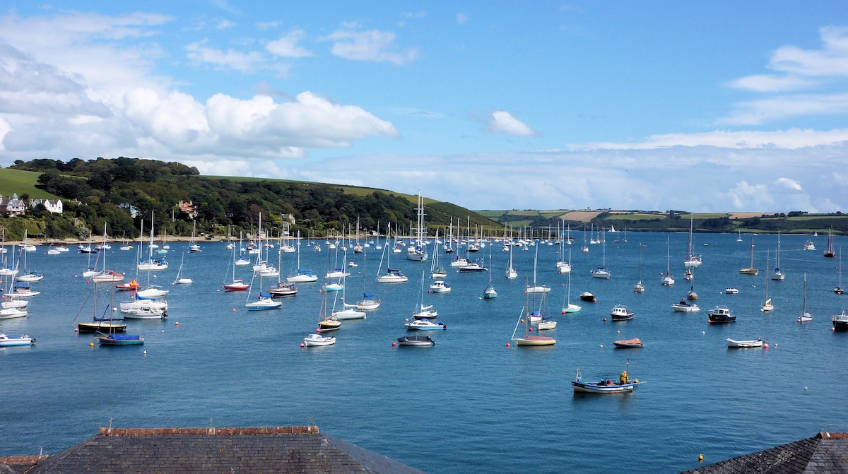 A scenic view of a harbor filled with numerous sailboats and fishing vessels, gently floating on calm blue waters under a partly cloudy sky. Lush green hills are visible in the background, enhancing the tranquil atmosphere of the coastal landscape.