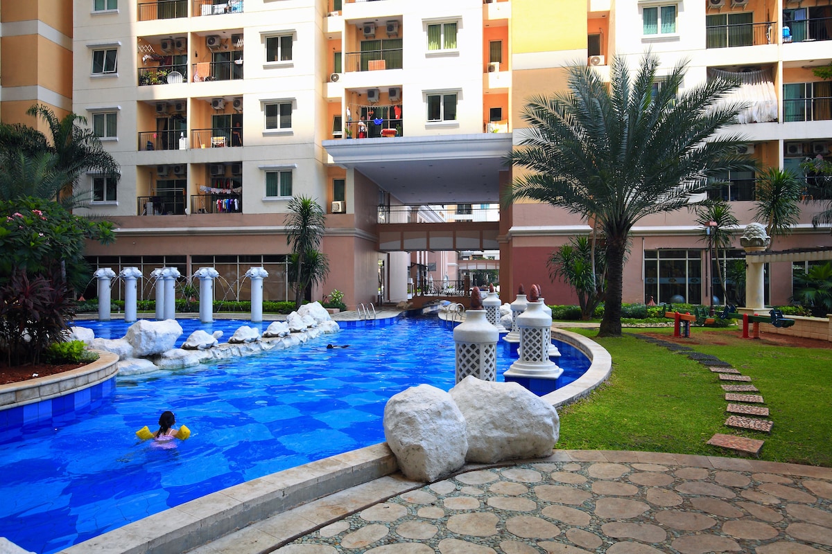 A large swimming pool features a vibrant blue surface surrounded by lush greenery and decorative rocks. Palm trees provide shade near the water, while a pathway of stones leads to seating areas. The building's entrance is visible in the background, with balconies overlooking the pool.