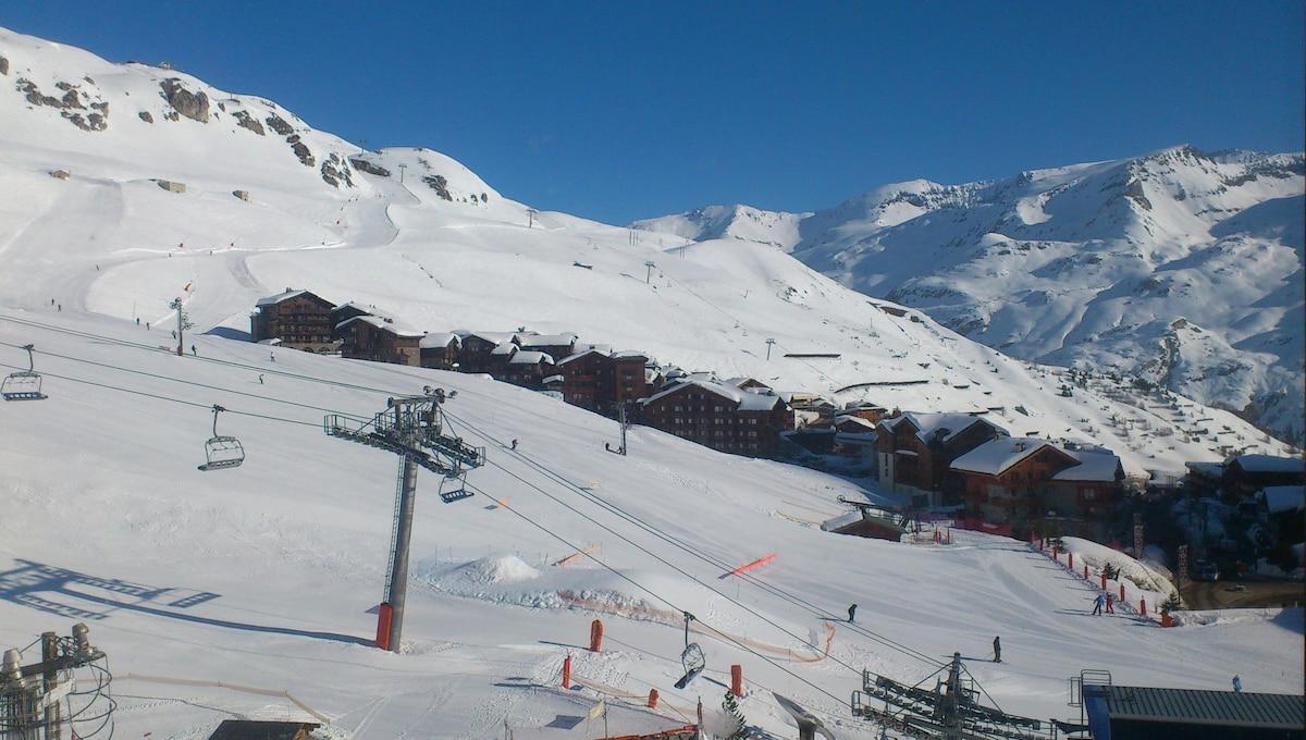 A panoramic view showcases a snowy landscape featuring ski slopes and multiple wooden chalets. Ski lifts are visible in the foreground, while mountains stretch across the horizon under a clear blue sky.