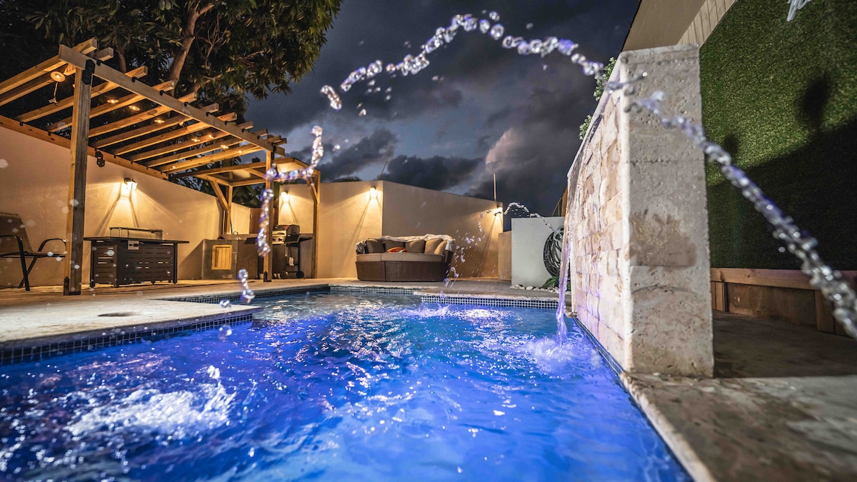 A private pool is highlighted in this modern outdoor space, featuring sparkling blue water and playful water jets. An inviting seating area is positioned nearby under a wooden pergola, softly illuminated by lights. Lush greenery lines the perimeter, enhancing the serene ambiance.