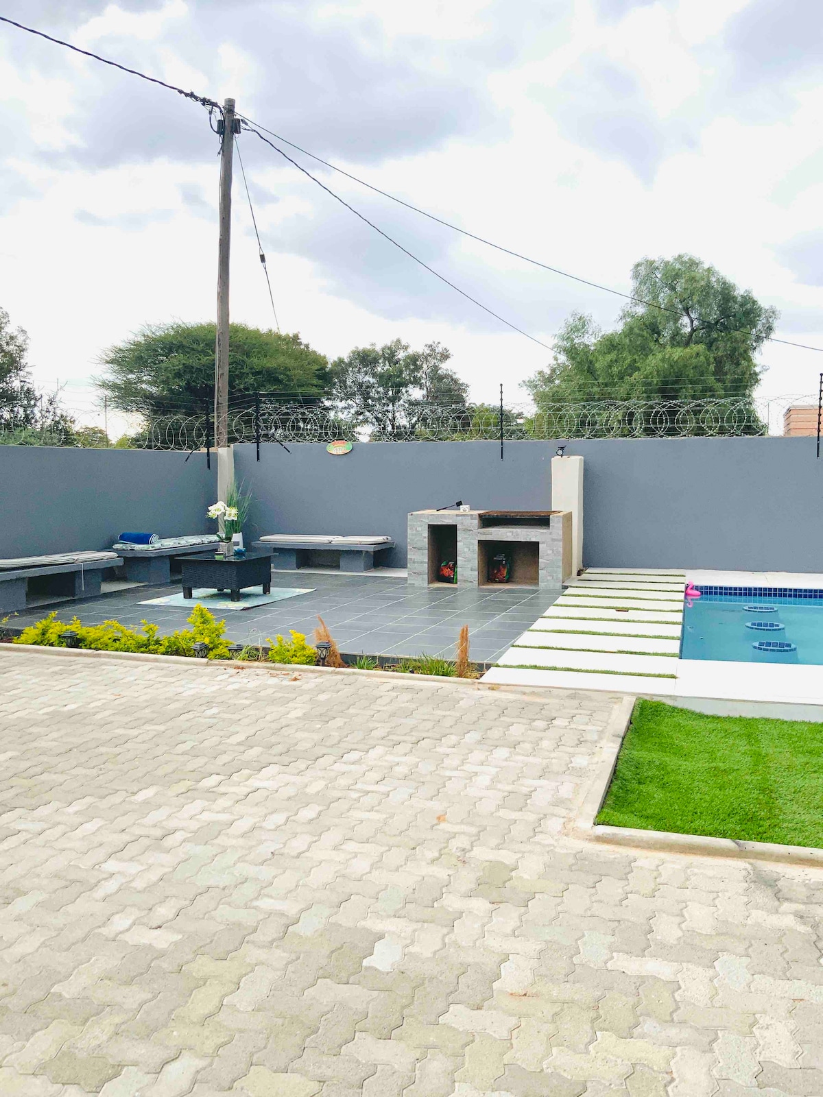An outdoor area features a sleek patio with seating arranged around a charcoal grill. A small pool reflects the surrounding greenery. The space is enclosed by a gray wall, with stepping stones leading across the manicured lawn.