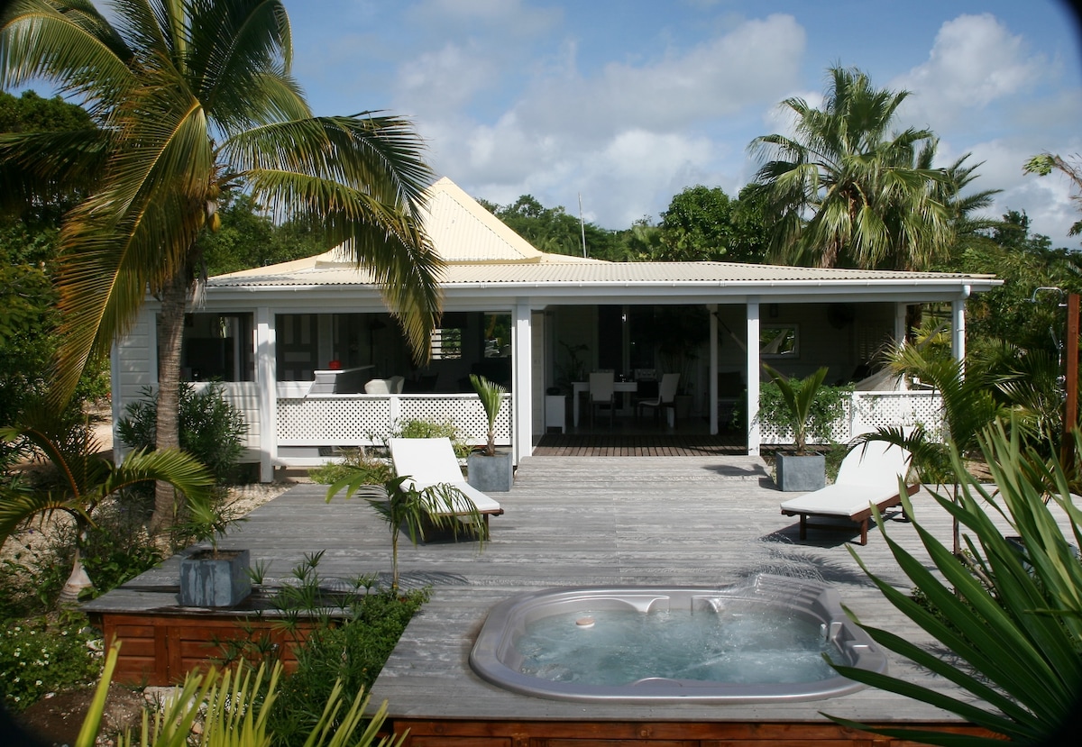 A guest house is set amidst lush greenery, featuring a spacious wooden deck. A hot tub is positioned on the side, accompanied by lounge chairs. The front opens to a shaded area, connecting to the interior living spaces. Palm trees add to the tropical ambiance.