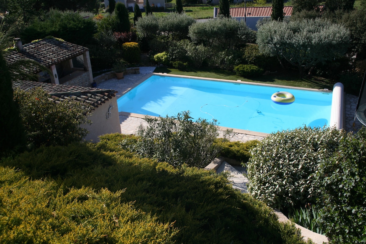 A sparkling blue swimming pool is framed by lush greenery and well-maintained landscaping. A relaxing pool float is visible on the water's surface. The surrounding area is enhanced by Mediterranean-style foliage and an outdoor terrace, offering a serene and inviting outdoor retreat.