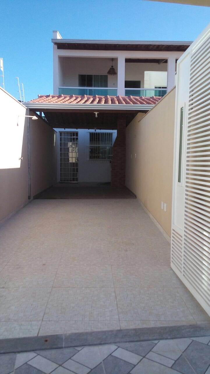 A covered area is visible, leading toward an entrance with a gated door in the background. The space features tiled flooring and walls, with natural light filtering in from the side. The roof is supported by columns, creating a shaded walkway.