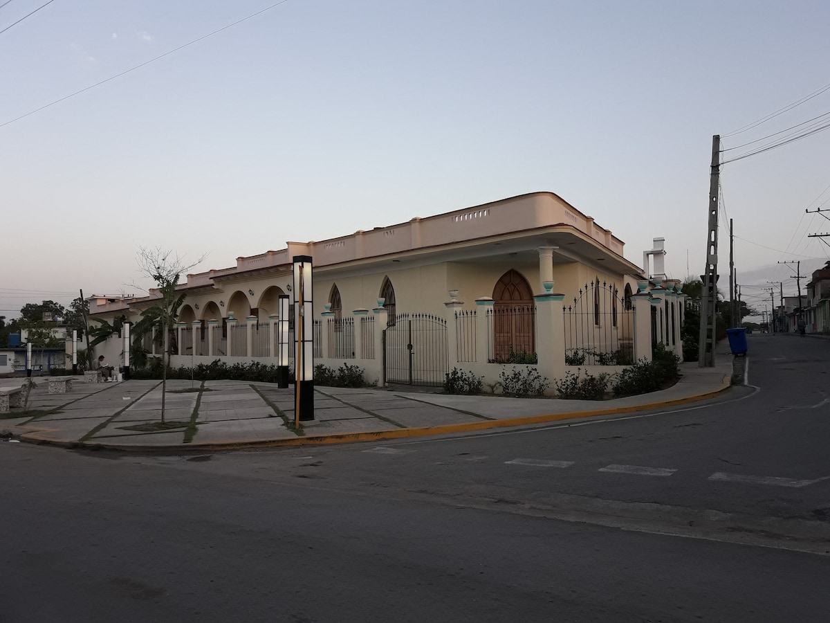A single-story building with an arched façade is situated on a corner lot. The exterior features cream-colored walls, detailed doorways, and a decorative fence. Surrounding pathways and landscaping enhance the inviting appearance, while street elements such as a lamppost provide additional context.