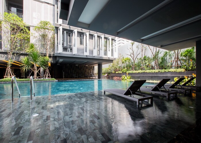 A serene swimming pool reflects surrounding greenery, with modern lounge chairs positioned along the water's edge. The area is shaded by a low-rise building, creating a peaceful atmosphere for relaxation and leisure.