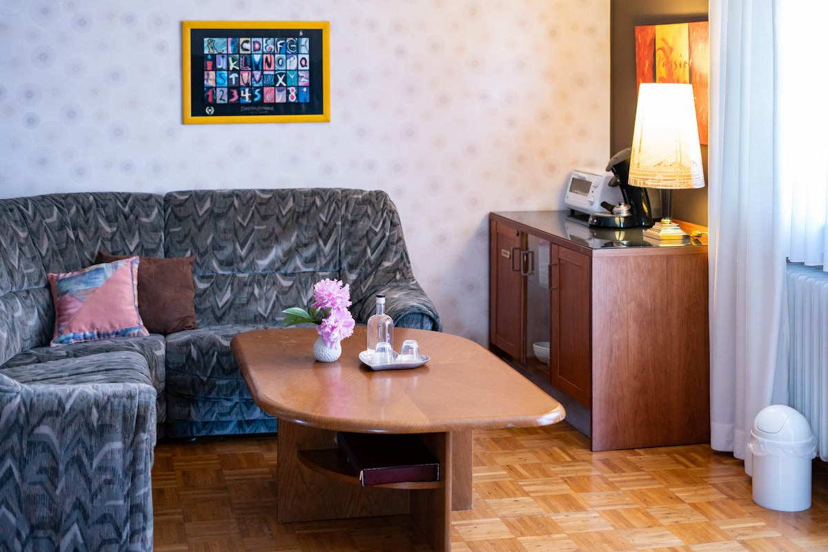 A cozy seating area features a plush sofa in earthy tones and a wooden coffee table adorned with a vase of pink flowers. A side table holds a lamp, illuminating the space. Textured wallpaper contrasts with framed artwork on the wall, adding visual interest.
