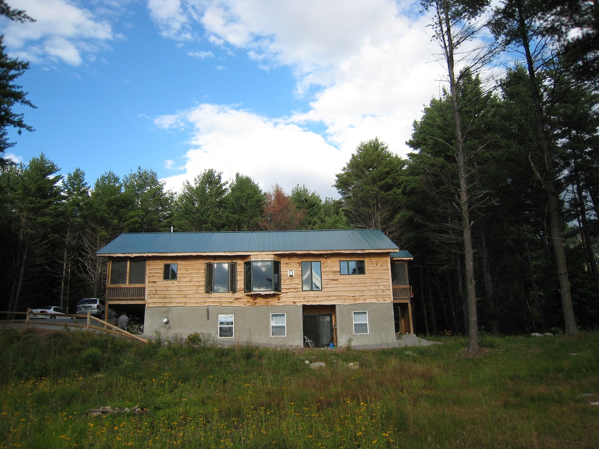 A spacious, two-story retreat is nestled amidst tall pines, featuring a combination of natural wood siding and a sturdy stone lower level. Large windows are framed by greenery, capturing the essence of the surrounding meadows and forests.