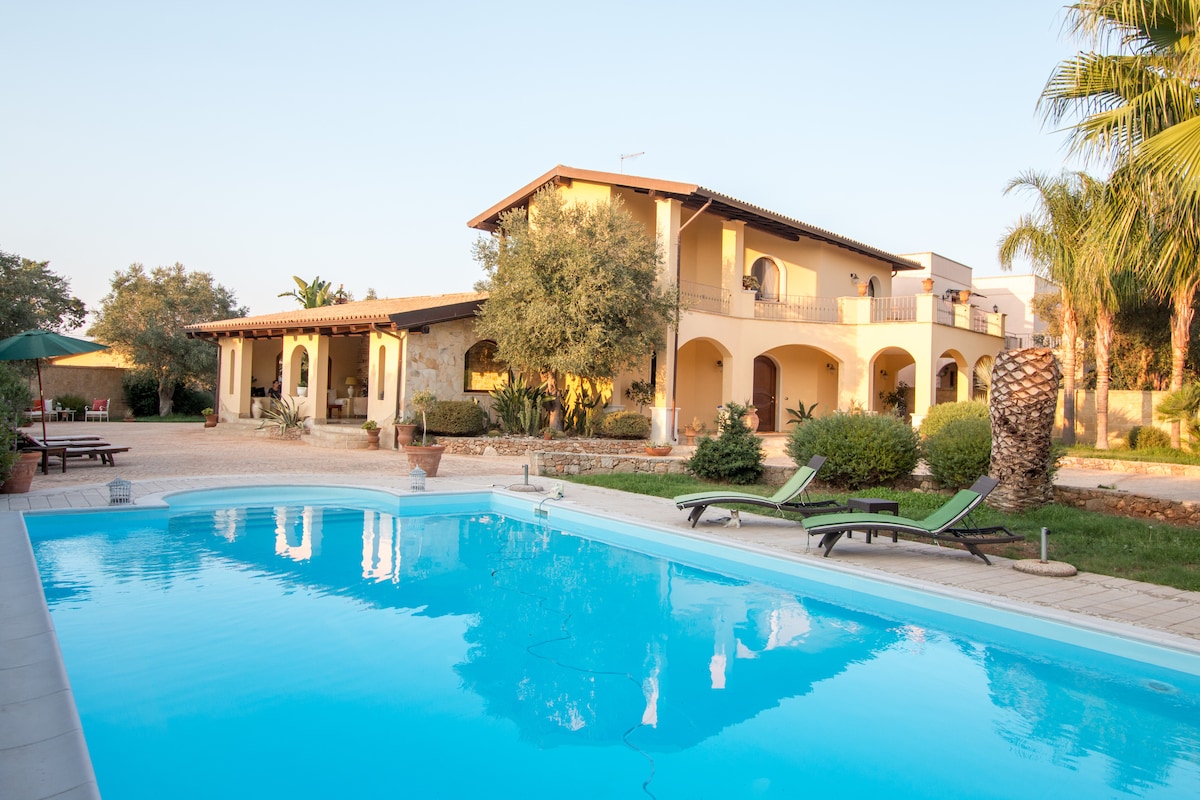 The exterior of the villa is shown, featuring a spacious pool surrounded by lush greenery and palm trees. Relaxing lounge chairs are positioned near the water. The two-story structure displays a warm color palette and multiple balconies, creating an inviting outdoor atmosphere.
