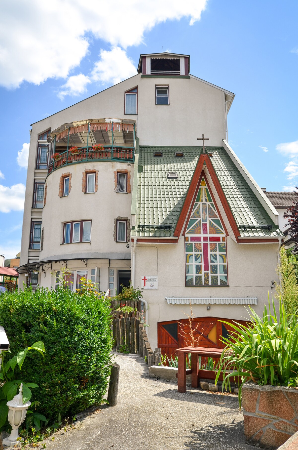 The building features a unique architectural design, showcasing a combination of angled roofs and a striking stained glass window. The exterior is painted in a light color, surrounded by greenery and a pathway leading to the entrance.