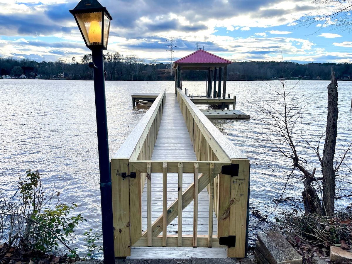 A wooden dock extends into Lake Wylie, featuring a safety gate at the entrance. A lamppost stands nearby, providing soft illumination. A covered lift is positioned at the dock's end, with serene water views and a backdrop of distant trees and clouds.