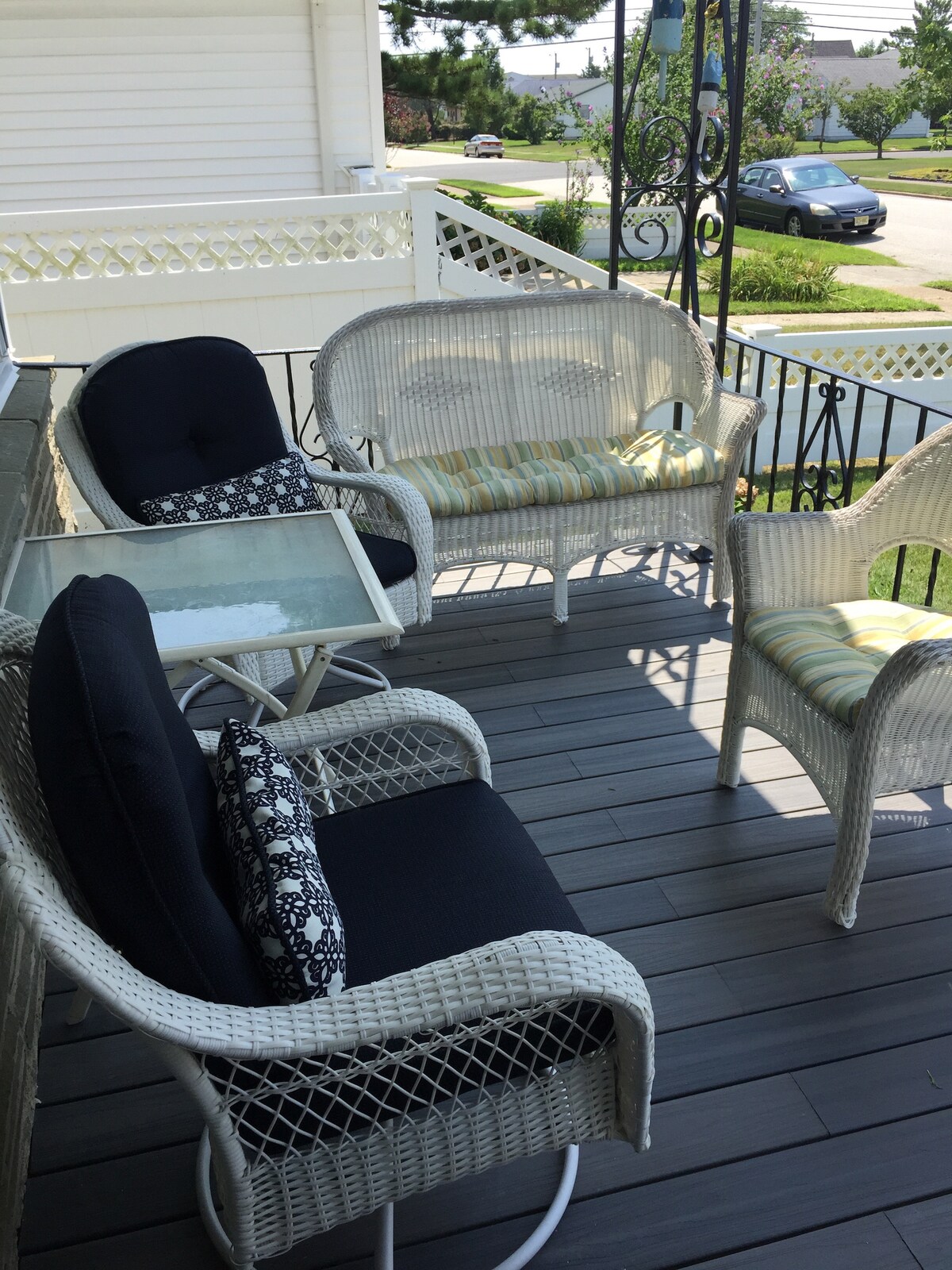 A cozy porch space is featured, furnished with four wicker chairs—two with cushions and a small glass table. The surrounding railing is visible, and the view of the quiet neighborhood is framed by greenery and paved streets.