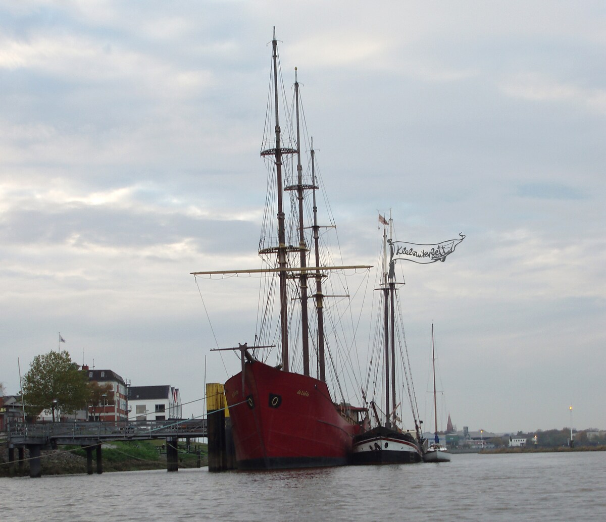 A traditional two-masted ship is anchored at a dock, displaying its striking red hull and multiple sails. Nearby boats are also visible, along with the riverside buildings and trees. A flag bearing the name of the ship is mounted on the mast, creating a nautical scene.